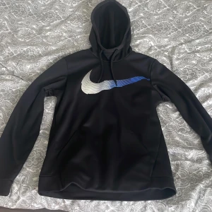 Svart Nike hoodie med blåvit logga - Säljer en svart hoodie från Nike med stor blåvit swoosh-logga på bröstet. Tröjan har huva med dragsko och är tillverkad i Dri-Fit material som andas. Perfekt för träning eller chill. Klassisk och stilren design. Hör av er vid frågor.