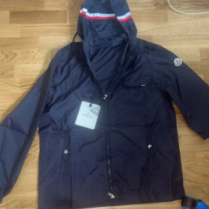 Mörkblå vindjacka från Moncler -  Äkta! Snygg mörkblå vindjacka från Moncler med huva och dragkedja framtill. Jackan har vita och röda detaljer på huvan samt Moncler-logga på ärmen. Perfekt för blåsiga dagar.