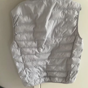 Ljusgrå dunväst med dragkedja - Snygg ljusgrå dunväst  med hög krage och dragkedja framtill. Västen har två fickor med dragkedja och ett diskret logomärke på bröstet. Perfekt för lager-på-lager under kyliga dagar.