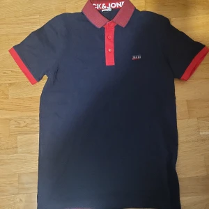 Mörkblå pikétröja från Jack & Jones - Säljer en mörkblå pikétröja från Jack & Jones med röda detaljer på krage, knappar och ärmslut. Klassisk passform och kortärmad modell, perfekt för en stilren look. Snygg logga på bröstet och bekvämt bomullsmaterial.