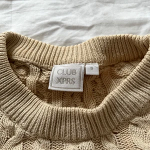 Beige kabelstickad tröja från CLUB XPRS - Säljer en beige kabelstickad tröja från CLUB XPRS. Tröjan har rund halsringning och ribbade muddar vid ärmslut och nederkant. Perfekt för lager-på-lager och en klassisk look.