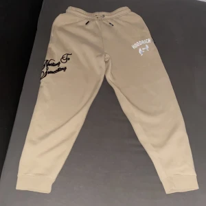 Beige mjukisbyxor från Hoodrich - Säljer ett par beige mjukisbyxor från Hoodrich med tryck av två vita duvor och logga på ena benet. Byxorna har resår i midjan och muddar vid bensluten. Snygg broderad textdetalj på sidan.