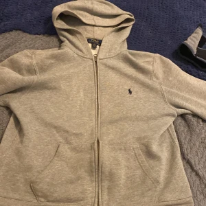 Grå hoodie från Polo Ralph Lauren - Säljer en klassisk grå hoodie från Polo Ralph Lauren med dragkedja och huva. Tröjan har två fickor framtill och den ikoniska broderade loggan på bröstet. Perfekt för en avslappnad stil.