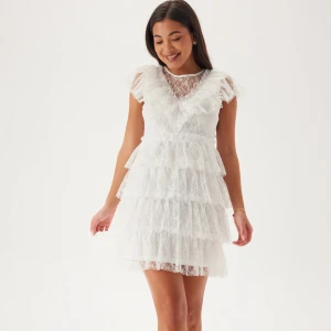 Vit spetsklänning med volanger - Superfin vit kortklänning med flera lager volanger och spetsdetaljer. Klänningen har v-ringning och är ärmlös med en romantisk känsla. Perfekt för sommarens alla tillfällen och ger en söt och trendig look.