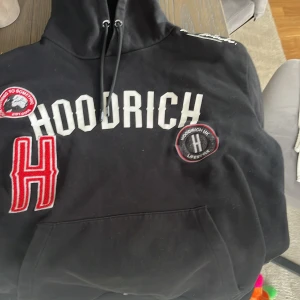 Svart hoodie från Hoodrich med patchar - Svart hoodie från Hoodrich i storlek M. Tröjan har stora vita bokstäver framtill, röda och svarta patchar samt huva med dragsko. Flera broderade detaljer och känguruficka framtill. Perfekt för en streetwear-look.