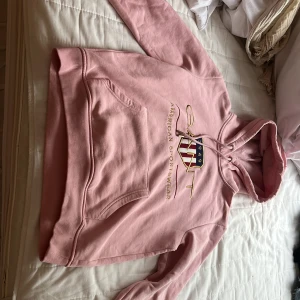Rosa hoodie från GANT - Säljer en rosa hoodie från GANT med broderad logga och text på bröstet. Tröjan har huva med dragsko och en stor magficka. Mjuk bomullskvalitet och ribbade muddar vid ärmslut och nederkant.