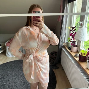 Ljus omlottklänning med knyt i midjan - Säljer en ljus kortklänning med krage och långa ärmar. Klänningen har omlottmodell och knyt i midjan, med ett mjukt mönster i pastelliga toner av vit och aprikos. Perfekt för vår och sommar!