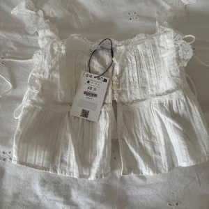 Topp  - Supersöt vit blus med spetsdetaljer och knappar framtill. Axelbandslös design med romantiska volanger och broderade detaljer. Perfekt för en somrig look!