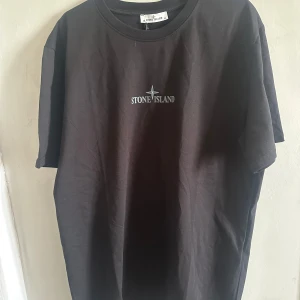 Svart t-shirt från Stone Island - Säljer en svart t-shirt från Stone Island med logga tryckt på bröstet och stort tryck på ryggen. Klassisk rund halsringning och normal passform. Perfekt för dig som gillar streetwear och vill ha något stilrent med tydlig branding.