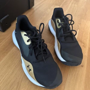 Svarta och guldiga sneakers/ basketskor från Under Armour - Snygga svarta sneakers med guldiga detaljer från Under Armour. Skorna har snörning, vit sula och rund tå. Perfekta för träning eller vardagsstil.