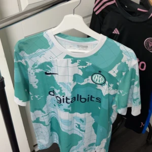 Inter Milan fotbollströja digitalbits - Säljer en Inter Milan fotbollströja med digitalbits sponsortryck. Tröjan är kortärmad och har ett unikt mönster i turkos, vit och inslag av svart. På ryggen står det KARIMINHO och nummer 77. Perfekt för fans av klubben!