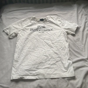 Vit t-shirt från Peak Performance - Vit t-shirt från Peak Performance med svart tryck på bröstet. Klassisk passform och rund halsringning. Perfekt basic-plagg med stilren design och mjukt bomullsmaterial.