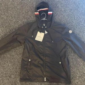 Svart vindjacka från Moncler med huva - Svart vindjacka från Moncler med huva och dragsko. Jackan har dragkedja framtill, två fickor med knappar och Moncler-logga på ärmen. NFC scan funkar, helt nytt oanvänd, säljer för den va för liten för mig, pris går att diskuteras.