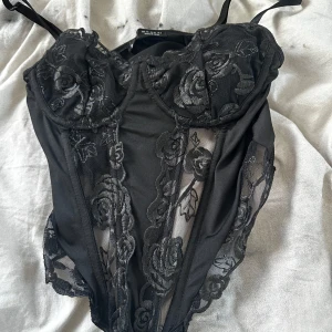 Svart spetskorsett med blommönster från Shein - Säljer en svart korsett med vackra spetsdetaljer och blommigt mönster från shein. Modellen har smala axelband, genomskinliga partier och markerade kupor för en snygg siluett. Perfekt för att lyfta vilken outfit som helst. ❤️