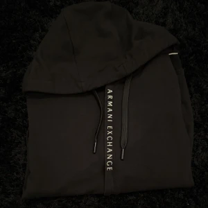 Svart hoodie från Armani Exchange - Säljer en svart hoodie från Armani Exchange med dragkedja framtill och huva. Tröjan har snören med logotypen tryckt längs dragkedjan och ribbade muddar vid ärmslut och nederkant. Perfekt för en stilren och avslappnad look.