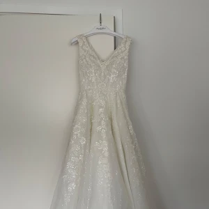 Brudklänning Vit glittrig långklänning med spetsdetaljer - Elegant vit brudklänning långklänning med v-ringning och breda axelband. Klänningen har glittrande paljetter och vackra spetsapplikationer över hela tyget. Perfekt för speciella tillfällen när du vill känna dig extra fin. Pris går att diskutera. Köpt på ivory and grace för 24 000kr. 