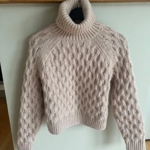 Beige stickad polotröja - Säljer en beige stickad polotröja med grovt kabelstickat mönster. Tröjan har hög polokrage och långa ärmar, perfekt för kyliga dagar. Den är mjuk och har en klassisk, tidlös look som passar till det mesta.
