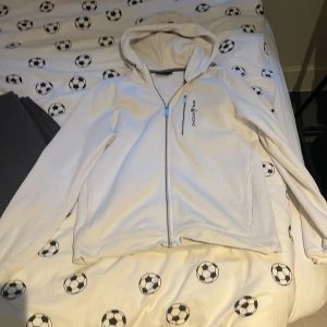 Vit hoodiejacka från Sail Racing - Vit hoodiejacka från Sail Racing med dragkedja framtill och en extra dragkedjeficka på bröstet. Jackan har huva och är långärmad, perfekt för en sportig och avslappnad stil. Inte använd så många gånger pris kan diskuteras 