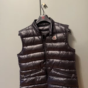 Svart dunväst från Moncler - Snygg svart dunväst från Moncler med hög krage och dragkedja framtill.passar 160-170. Dragkedjan är lite trasig men går att dra upp med tålamod(även därför det är bra pris) Västen har två fickor med dragkedja och Moncler-logga på bröstet. Perfekt för sena kvällar på sommaren!🥳🍾frågor eller funderingar hör bara av er!