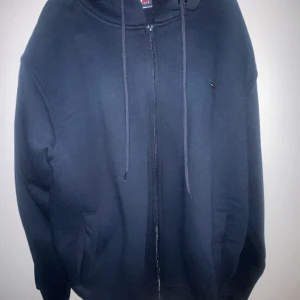 Mörkblå hoodie med dragkedja från Tommy Hilfiger - Säljer en mörkblå hoodie från Tommy Hilfiger med dragkedja framtill och justerbar huva. Tröjan har två fickor och diskret logga på bröstet samt ärmen. Perfekt för en avslappnad stil. Hör av er vid funderingar😁