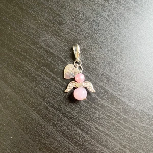 Rosa ängelberlock med vingar💕 - Söt berlock i form av en ängel med två rosa pärlor och silvriga vingar. Fästet är i silverfärg och det finns en liten bricka med texten 'Made with love'. Perfekt som accessoar till halsband eller armband.💕
