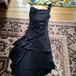 Svart asymmetrisk klänning med volang - Säljer en svart långklänning med asymmetrisk skärning och volangdetaljer. Klänningen har smala axelband och en axelbandslös sida, vilket ger en unik look. Perfekt för dig som vill sticka ut med en elegant och modern stil.