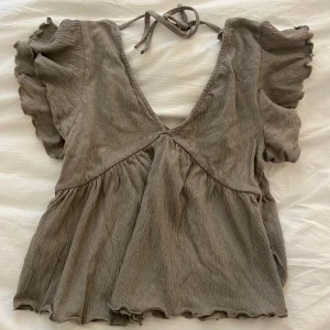 Beige volangtopp - Supersöt beige topp från Lager 157 med volangärmar och djup v-ringning. Toppen har knyt i nacken och är tillverkad i ett mjukt, ribbat material. Perfekt för dig som gillar romantiska detaljer och en luftig känsla.