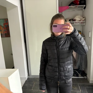 Svart Moncler pufferjacka - Säljer en svart pufferjacka från Moncler med klassisk logga på ärmen. Jackan har dragkedja framtill, två fickor med knappar och en stilren, quiltad design. Perfekt för dig som vill ha en lätt och snygg jacka med streetkänsla. Dåligt med dun därav priset