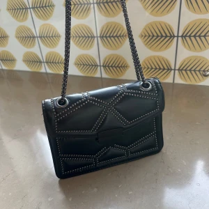 Svart axelväska med nitar och kedja - Snygg svart axelväska i skinn med coolt geometriskt mönster av silvriga nitar. Väskan har en kedjerem och ett stort fack med innerficka med dragkedja. Perfekt storlek för det viktigaste och ger en ”put together” vibe till din outfit. Perfekt inför väskflrbudet som tagits bort för festivalerna/konserterna i sommar.