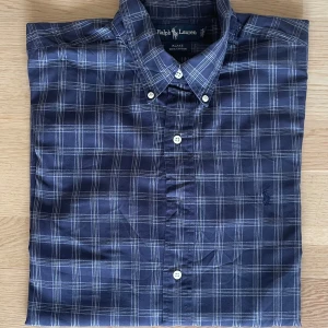 Blårutig skjorta från Ralph Lauren XL - Snygg blårutig skjorta från Ralph Lauren i 100% bomull. Klassisk button-down krage, vita knappar och diskret broderad logga på bröstet. Perfekt för dig som gillar en tidlös och stilren look med lite preppy känsla.