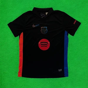 Svart FC Barcelona Nike fotbollströja - Snygg svart FC Barcelona fotbollströja från Nike med klubbmärke och Nike-logga på bröstet. Spotify-logga i rött framtill och röda samt blå paneler på sidorna. Tillverkad i lätt och ventilerande material, perfekt för match eller träning.