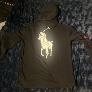 Svart hoodie från Polo Ralph Lauren - Svart hoodie från Polo Ralph Lauren med stor vit polospelare på ryggen och liten logga på bröstet. Snygg röd Ralph Lauren-signatur längs ena ärmen. Långärmad modell med huva och snörning, tillverkad i mjuk bomull. Stor Ralph Lauren logga på ryggen, svart färgad tvättas såklart innan den skickas 
