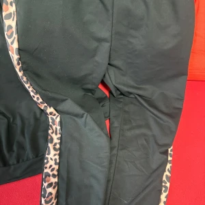 Svarta mjukisbyxor med leoparddetalj XL - Svarta mjukisbyxor med bred resår i midjan och snygga leopardmönstrade ränder längs sidorna. Byxorna har en loose passform och är perfekta för en chill och trendig look. Tillverkade i mjukt material som känns skönt mot huden.