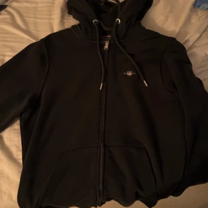 Svart hoodie från Gant med dragkedja - Svart hoodie från Gant med huva och snörning, dragkedja framtill och två fickor. Klassisk logga broderad på bröstet. Perfekt för en chill och stilren look.