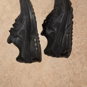 Svarta Nike Air Max sneakers - Snygga helsvarta Nike Air Max sneakers med klassisk synlig luftbubbla i sulan. Skorna har mesh och syntetmaterial för en sportig look, samt snörning och rund tå. Perfekta för dig som gillar streetstyle och vill ha bekväma skor med ikonisk design.