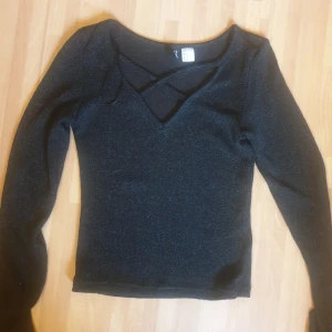 Svart glittrig topp från Divided - Svart långärmad topp från Divided med glittrigt tyg och snygg v-ringning med korsade band framtill. Toppens material har en lätt skimrande effekt som gör den extra iögonfallande. Perfekt för dig som gillar att sticka ut lite extra.