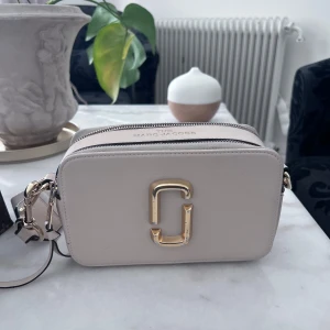 Beige axelväska från Marc Jacobs - Säljer en stilren beige axelväska från Marc Jacobs med guldigt logospänne framtill. Väskan har två dragkedjor upptill och en bred axelrem med Marc Jacobs-tryck. Tillverkad i skinn och har en boxig, modern form som passar till många outfits.