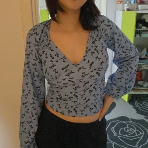 Ljusblå blommig omlottopp med öppen rygg - Supersöt ljusblå omlottopp med blommigt mönster och långa puffärmar. Toppen har v-ringning framtill och en snygg öppen rygg med knytband. Croppad passform som ger en trendig vibe, perfekt till jeans eller kjol.