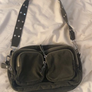 Grön axelväska med nitar och dragkedjor - Cool grön axelväska med bred axelrem dekorerad med silvriga nitar. Väskan har två framfickor med dragkedjor och är gjord i mjukt tygmaterial. Perfekt för att få plats med allt du behöver och ändå se stylish ut.