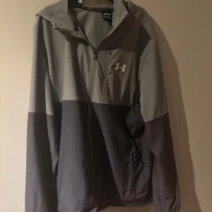 Under armour jacka - Snygg tvåfärgad vindjacka från Under Armour i ljusgrått och mörkgrått. Jackan har huva, dragkedja framtill och en bröstficka med dragkedja. Perfekt för blåsiga dagar och sportiga outfits. Priset kan diskuteras 