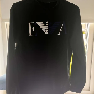 Svart sweatshirt från Emporio Armani - Snygg svart sweatshirt från Emporio Armani med stort EVA-logotryck i vitt och mörkblått framtill. Tröjan har rund halsringning och långa ärmar, perfekt för en stilren och trendig look.