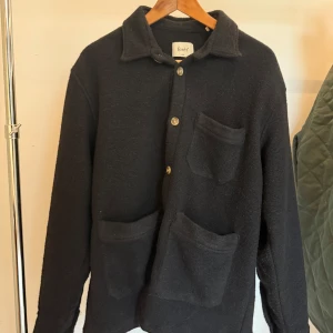 Svart overshirt från Forét i ullmix - Stilren svart overshirt från Forét med tre stora fickor framtill och klassisk krage. Skjortan är tillverkad i en mjuk ull- och polyestermix och har knäppning med knappar. Perfekt lager-på-lager-plagg med avslappnad passform.