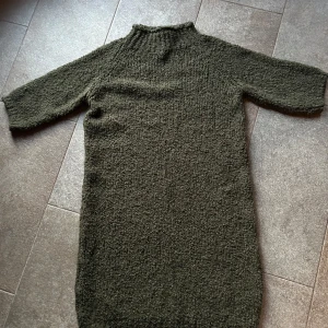 Olivgrön stickad klänning Marta - Mysig olivgrön stickad klänning från Marta med halvpolokrage och trekvartsärmar. Klänningen har en rak passform och är tillverkad i ett grovstickat material som känns både trendigt och bekvämt. Perfekt för lager-på-lager under kyliga dagar.
