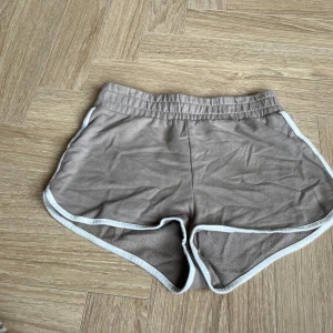 Beige frottéshorts från FB Sister - Snygga beige shorts från FB Sister i mjuk frotté med vita kanter. De har elastisk midja och sportig retrostil, perfekta för varma dagar eller chill hemma. Modellen är loose och har rundade benslut för en avslappnad vibe.