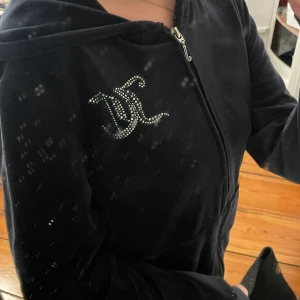 Juicy Couture mörkblå velourjacka - Svart velourjacka från Juicy Couture med glittrande strasstenar som bildar loggan på bröstet och stor Juicy Couture-text på ryggen. Jackan har dragkedja framtill och huva. en del av strassstenarna har åkt av på både framsidan o baksidan men det märks inte så tydligt, tyvärr åker det lätt av i tvätten. jag säljer tröjan pga att den inte kommer till användning. tröjan köptes från kidsbrandstore