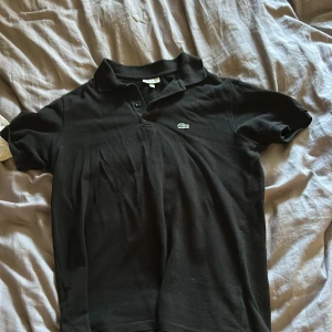 Svart pikétröja från Lacoste - Klassisk svart pikétröja från Lacoste med den ikoniska krokodilloggan på bröstet. Tröjan har korta ärmar, krage och knäppning framtill. Tillverkad i mjuk bomull som är skön mot huden. Perfekt för en clean och stilren look. Säljer pga att den bara hänger i garderoben 