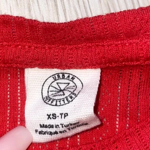 Röd ribbad topp från Urban Outfitters - Snygg röd topp från Urban Outfitters i ribbad bomull med korta ärmar och vågig kant vid ärmslut och nederkant. Toppen har en figurnära passform och diskret hålmönster med hjärtan i tyget. Riktigt fin tröja.