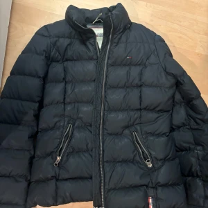 Svart pufferjacka från Hilfiger Denim - Säljer en svart pufferjacka från Hilfiger Denim med quiltad design och två dragkedjeförsedda fickor. Jackan har hög krage och klassisk logga på bröstet. Perfekt för kalla dagar och riktigt snygg till streetwear-stilen.