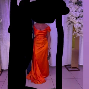 Lång orange satinklänning med drapering - Snygg långklänning i starkt orange satin med draperad framsida och lätt v-ringad hals. Klänningen har korta ärmar och markerad midja, samt en elegant slits på sidan. Perfekt för dig som vill sticka ut med färg och glans. Nypris:2800kr jag kan självklart skicka bild på hur den sitter på vid intresse storlek 40 men passar även 38
