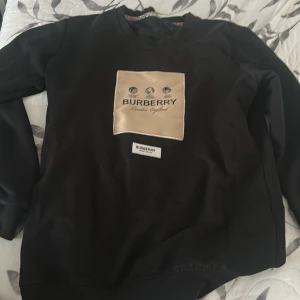 Svart Burberry sweatshirt med beige patch - Svart sweatshirt från Burberry med en stor beige patch på bröstet där loggan och texten 'London, England' syns. Tröjan har rund halsringning och långa ärmar. Perfekt för dig som gillar stilrena designer och exklusiva märken.